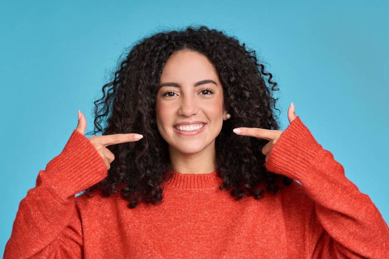 Smiling woman with curly dark hair wearing red sweater against blue background, pointing at her teeth.