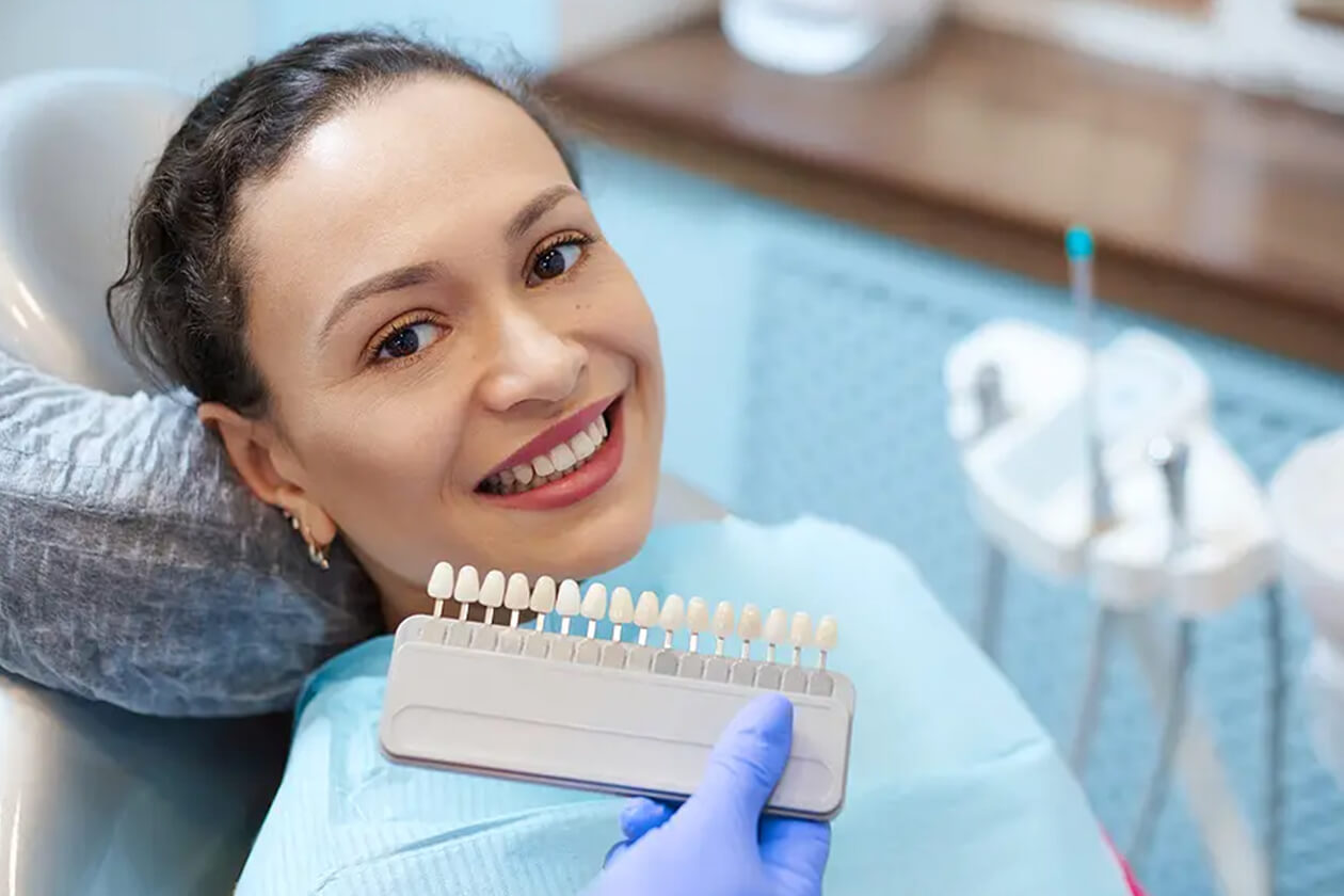 Woman smiling at dental office holding tooth shade guide for color matching during examination.