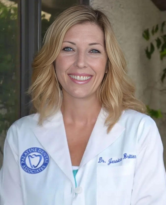 Female dentist in white coat with blue logo smiling at camera in dental office setting.