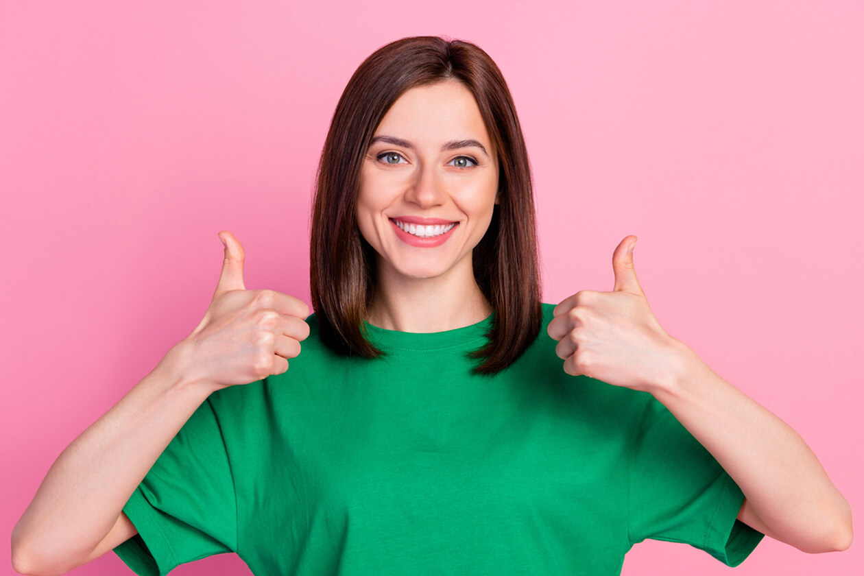 Woman in green shirt giving double thumbs up against pink background, smiling at camera
