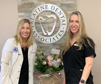 Two dental professionals smiling in front of Palatine Dental Associates sign with flowers.