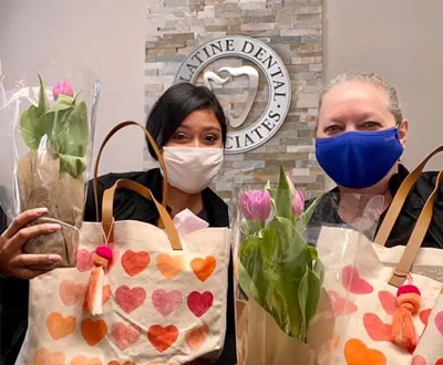 Two people wearing masks hold Valentine's Day gift bags in a dental office setting.