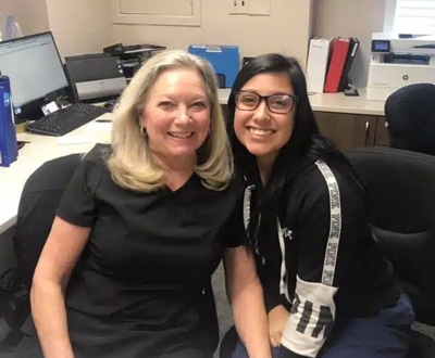 Two women smiling at camera in dental office with desk and computer equipment visible in background.