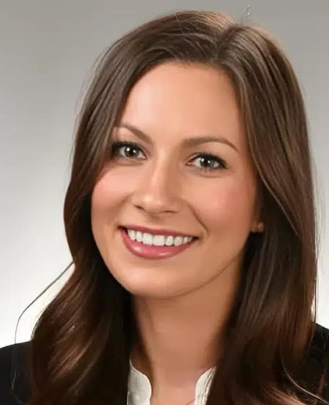 Professional headshot of smiling woman with long brown hair in business attire against neutral background.