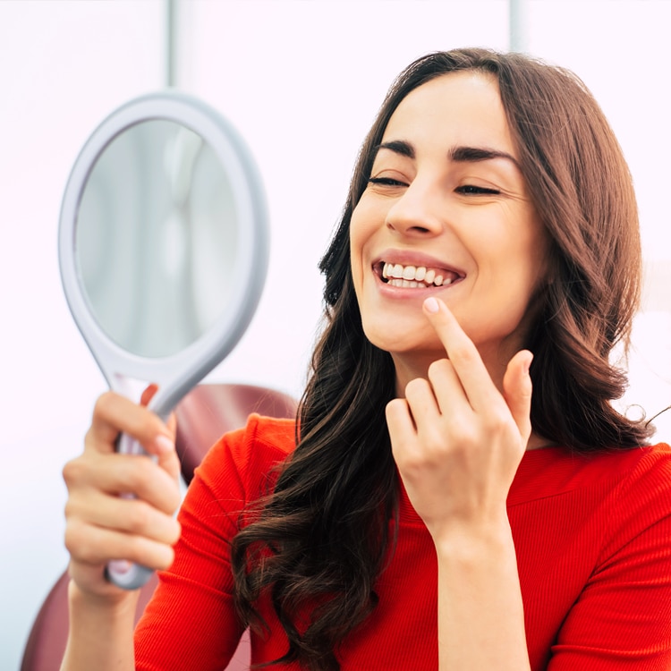 Woman in red shirt holding mirror, smiling while touching her face in dental office setting.