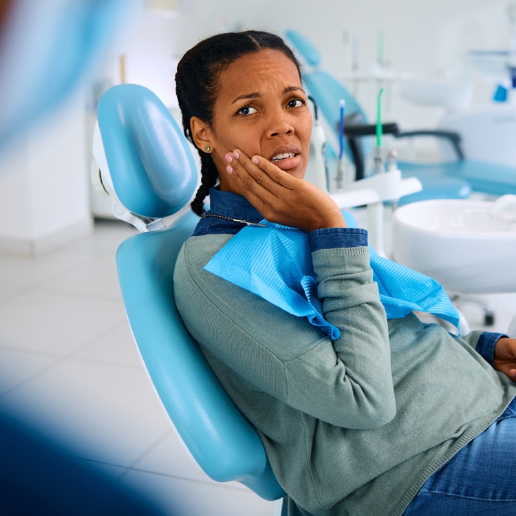 Woman in dental chair touching her cheek, appearing anxious or uncomfortable in dentist office.