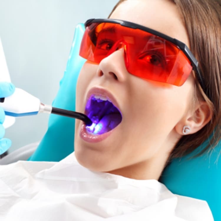 Patient wearing protective red glasses receives dental treatment with blue UV light in dental office.