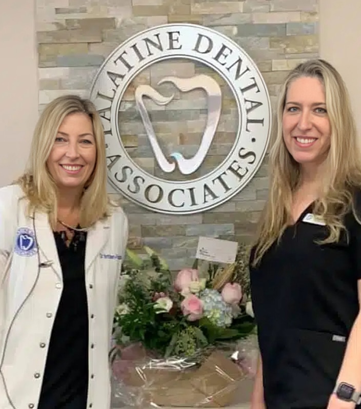 Two women smiling at Palatine Dental Associates office with logo and floral arrangement visible.