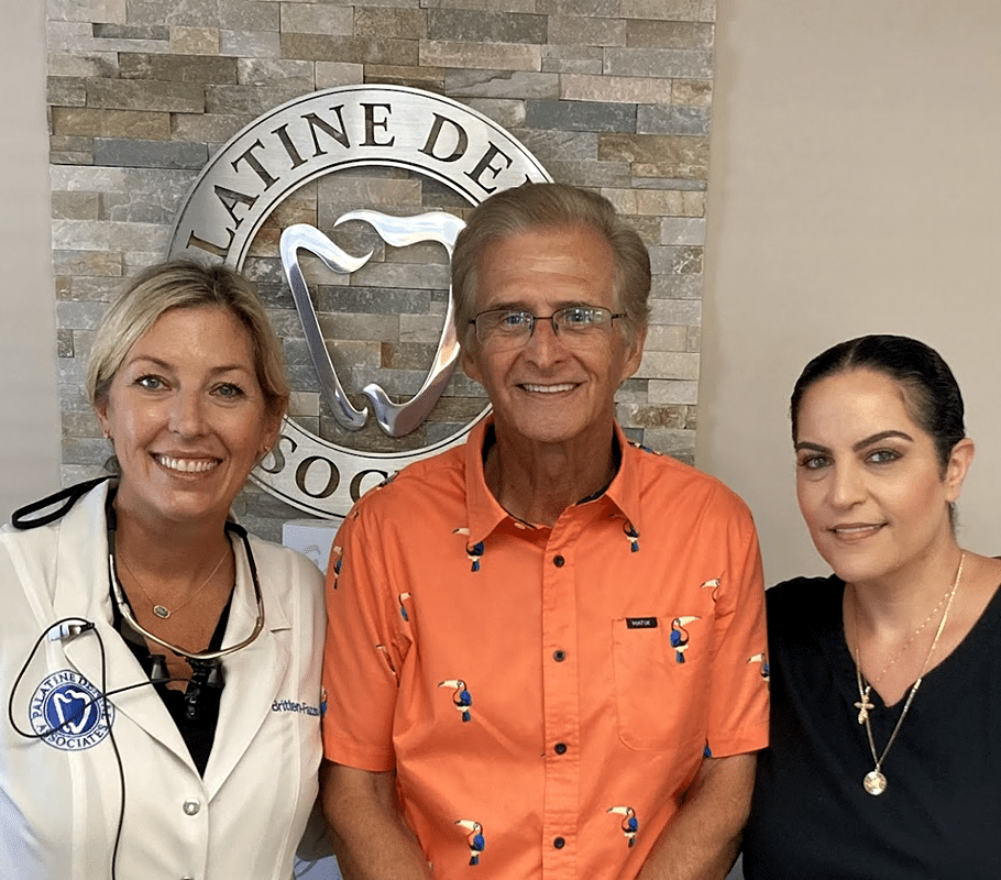 Three dental professionals posing together in front of Palatine Dental Associates logo and brick wall.