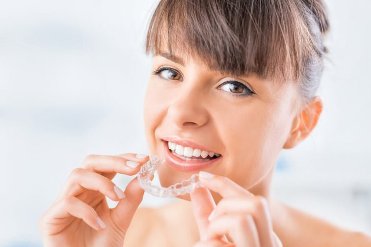 Young woman smiling while holding clear dental aligners in bright, clean dental office setting.
