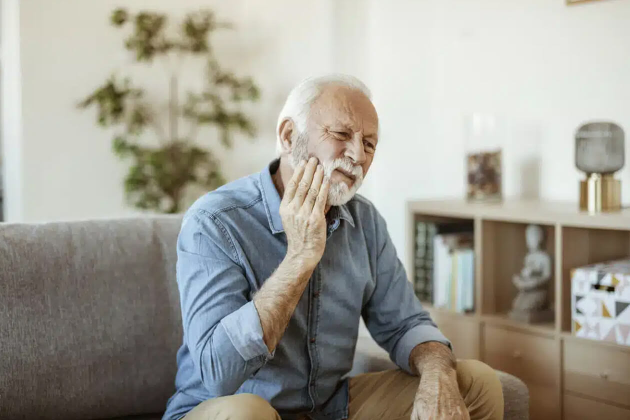 Senior man touching his cheek in pain while sitting on couch at home.
