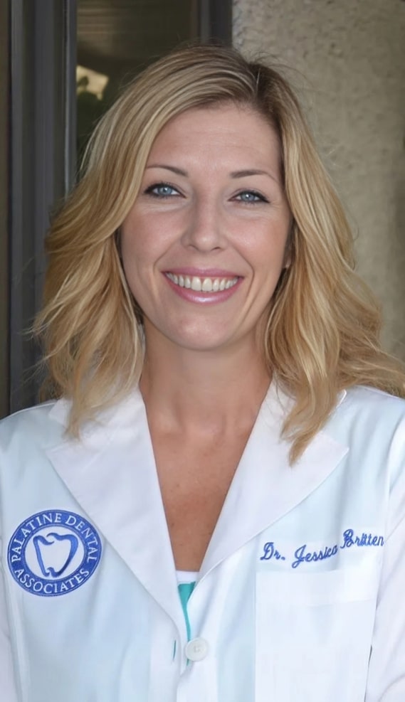 Female dentist in white coat with credentials smiling at camera in dental office setting.