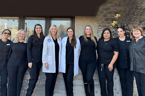 Dental team of twelve professionals standing together in front of clinic entrance wearing dark uniforms and white coat.