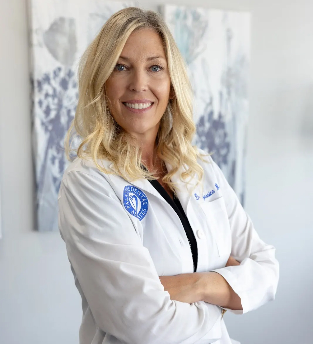 Professional female dentist in white coat with arms crossed, smiling at camera in clinical setting.
