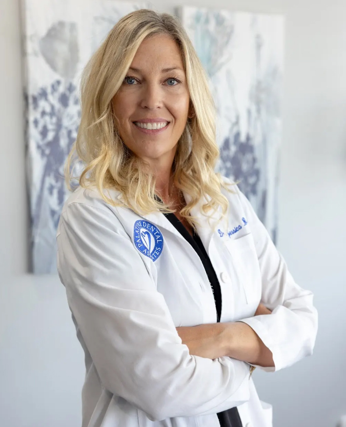Female dentist in white coat with arms crossed, smiling at camera in clinical office setting.