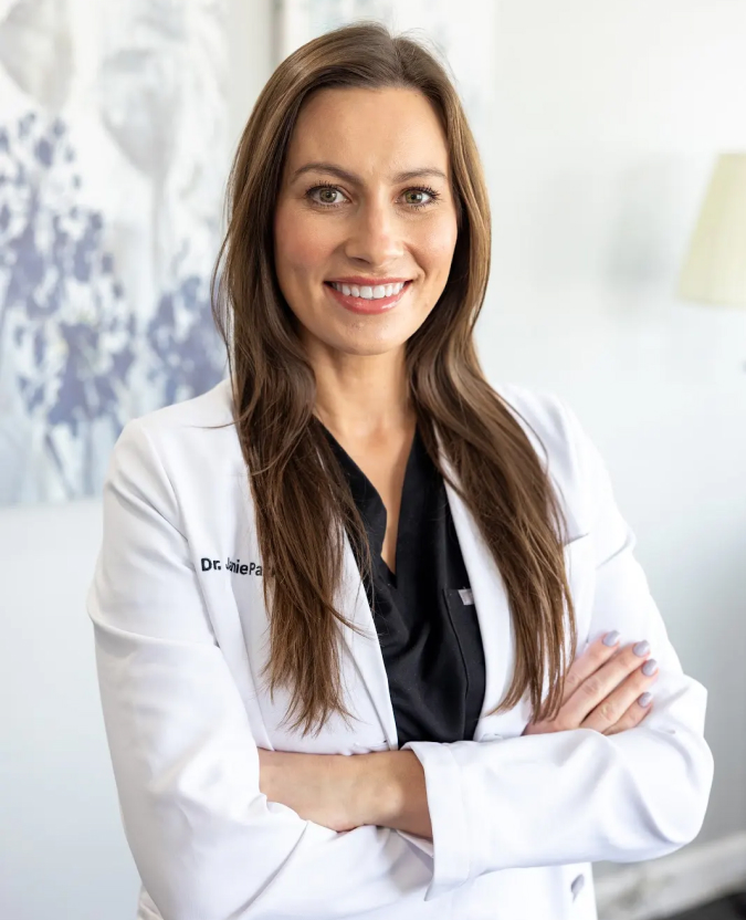 Professional female dentist in white coat smiling at camera with arms crossed in clinical office setting.