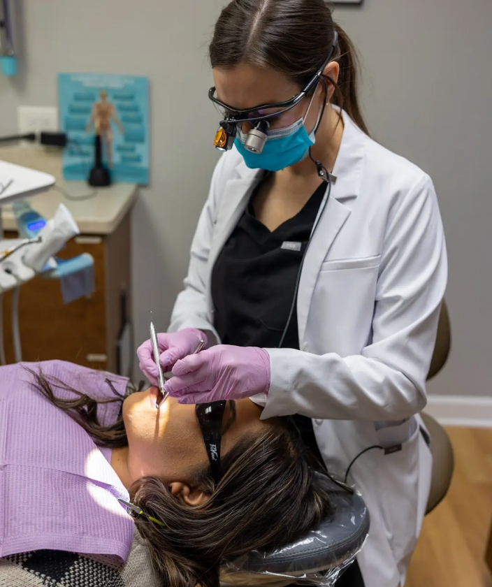 Dentist in white coat and protective gear examining patient's teeth in clinical setting.