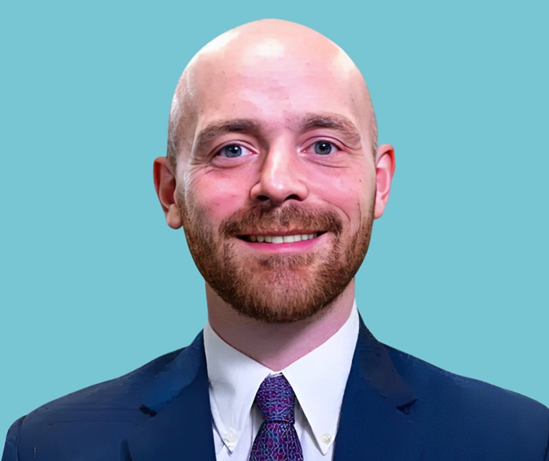 Professional headshot of smiling man in dark suit and purple tie against blue background.