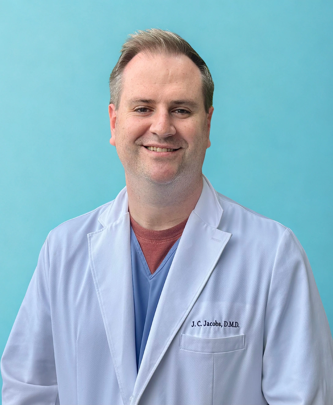 Male dentist in white coat and blue scrubs smiling against turquoise background.