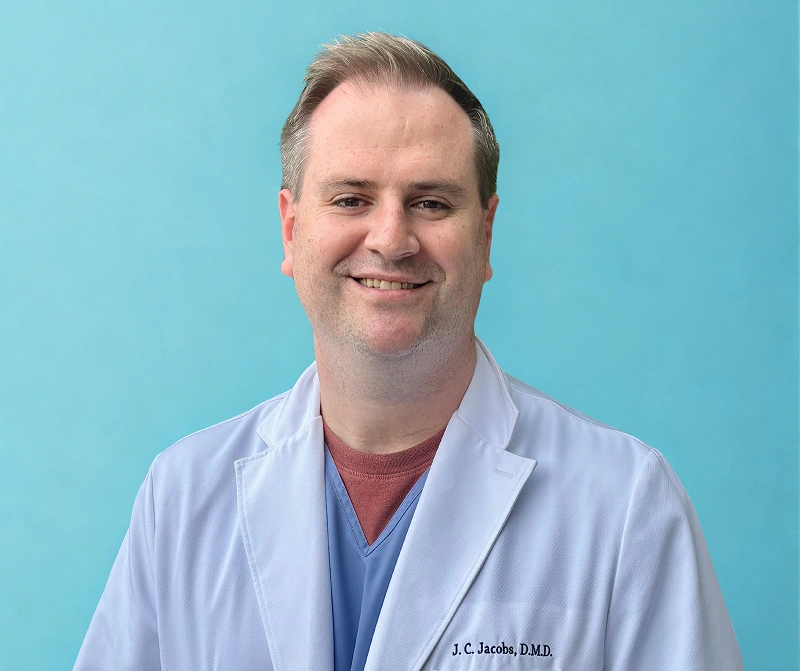 Male dentist in light blue coat smiling at camera against blue background.